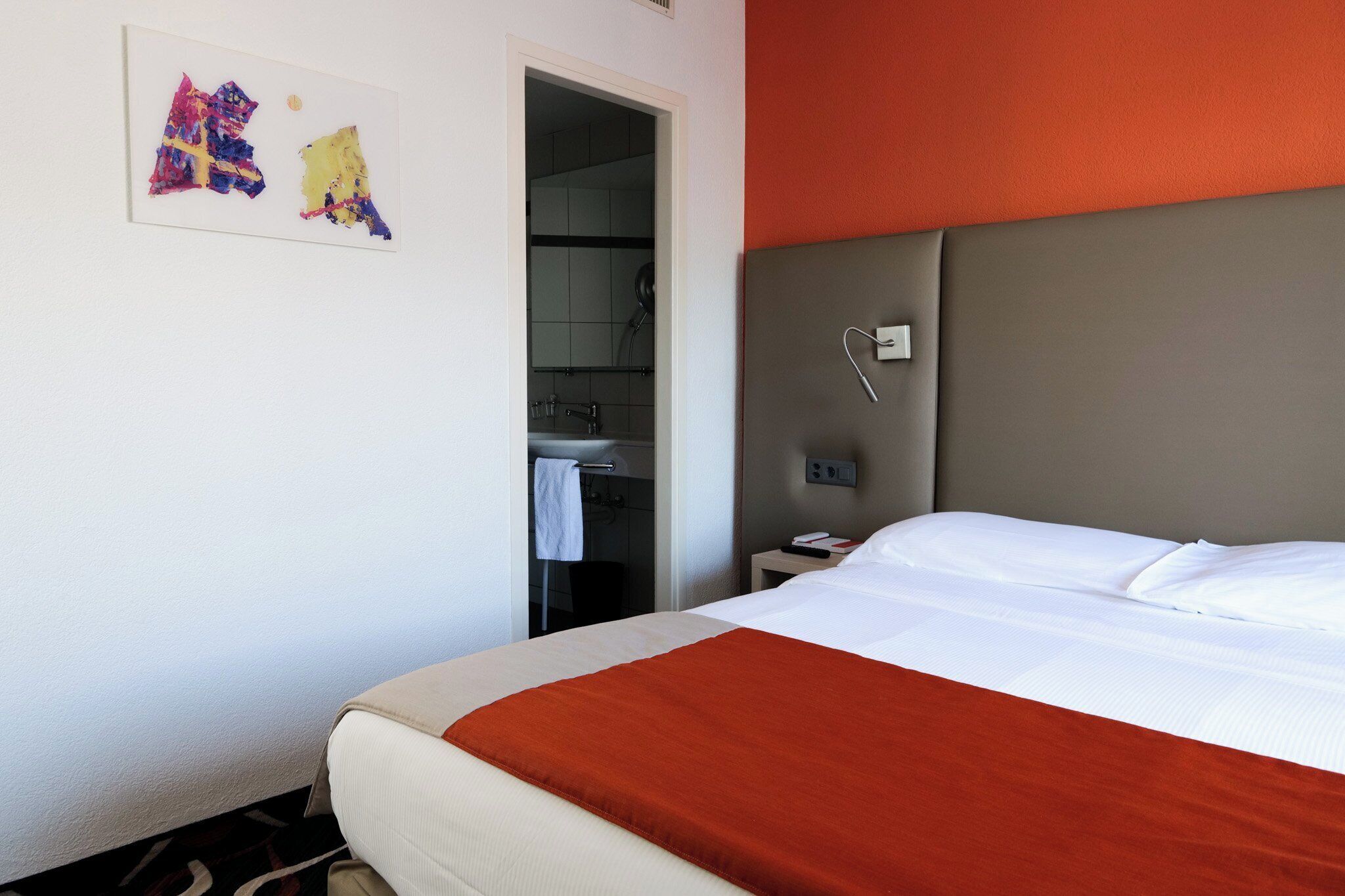 A hotel room with a bed, a nightstand, a wall-mounted light, and a door leading to a bathroom.