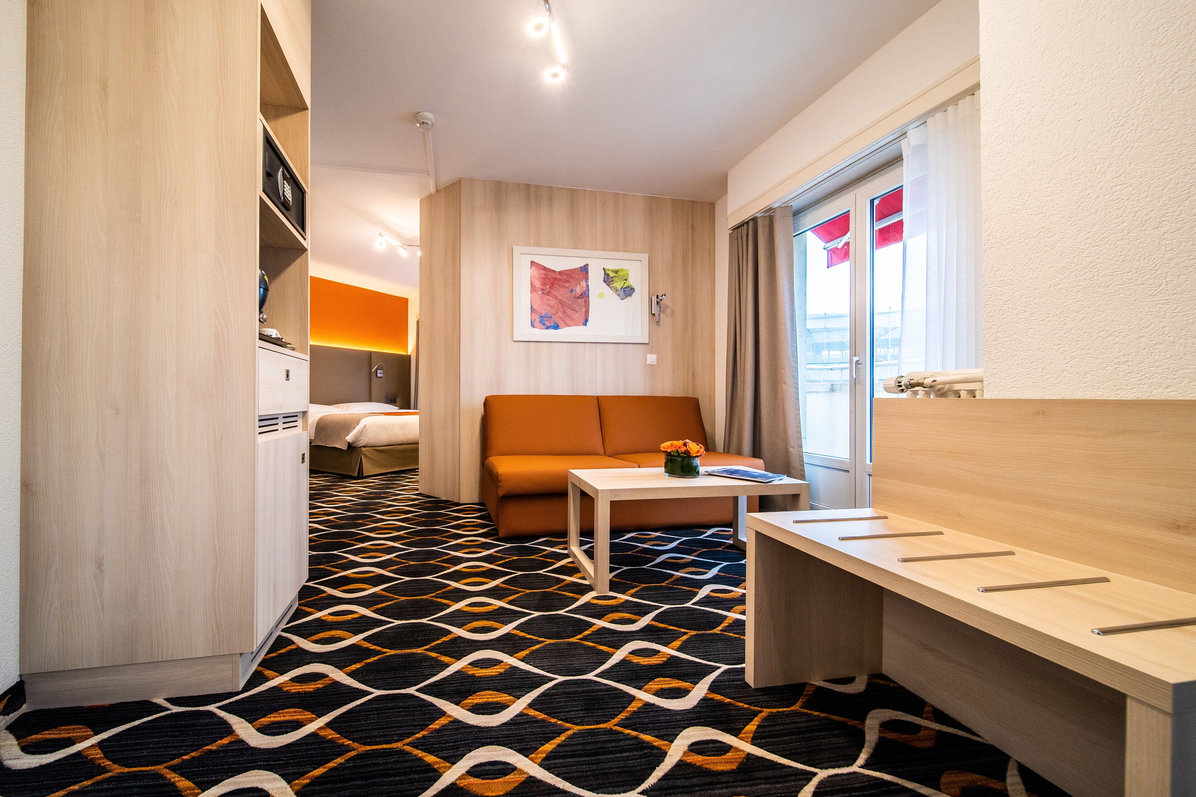 Ett modernt hotellrum med ett mönstrat golv, en soffa, ett litet bord med en vas, ett skrivbord i trä och en säng i bakgrunden.