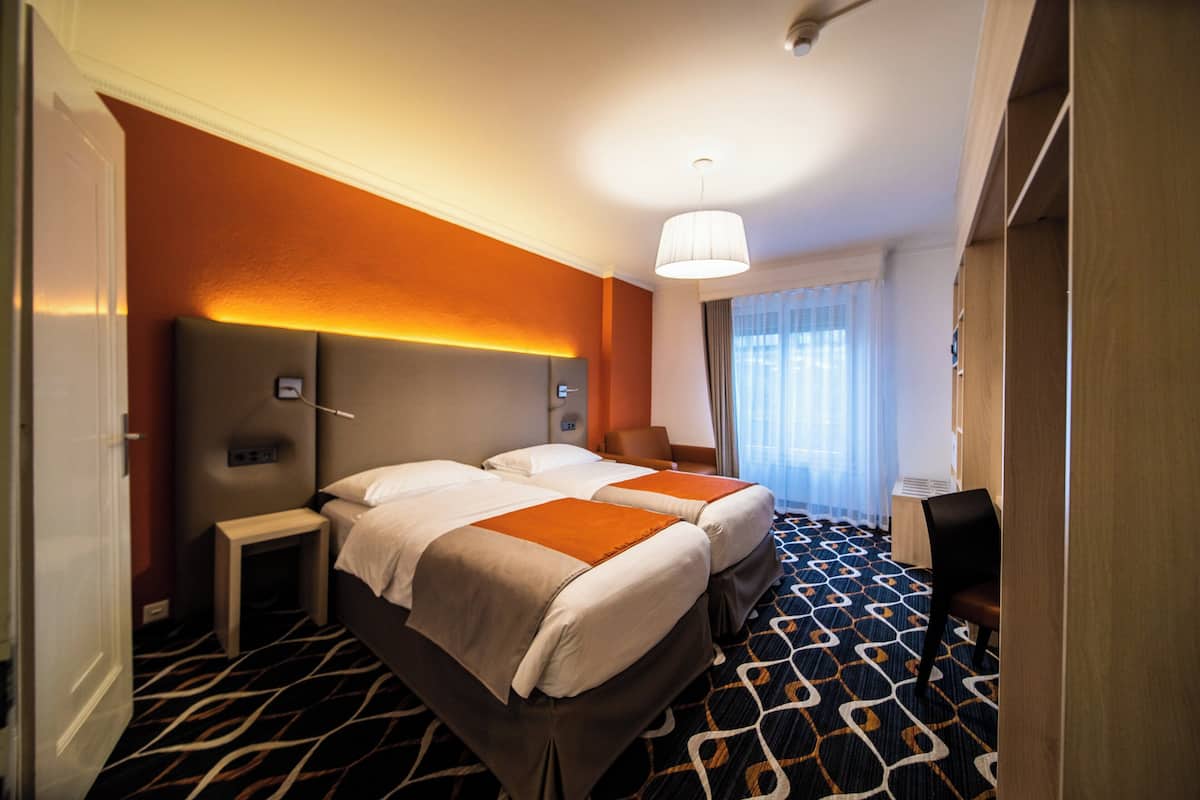 A hotel room with two beds, a bedside table, a chair, a desk, and a large window with curtains.