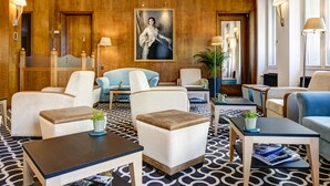 Un vestíbulo con alfombra estampada, varias zonas de estar que incluyen sillones y sofás, un cuadro central y lámparas fijadas a la pared.
