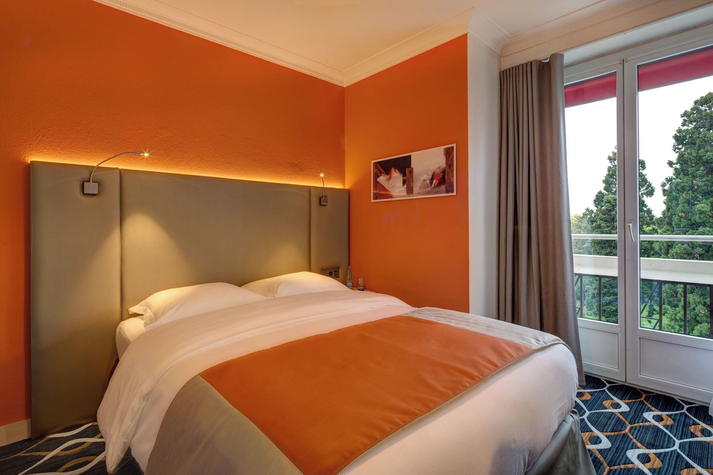 A hotel room with an orange wall, a bed with white and orange bedding, a headboard with integrated lighting, a window with curtains, and a view of greenery outside.