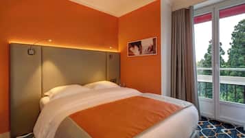 A hotel room with an orange wall, a bed with white and orange bedding, a headboard with integrated lighting, a window with curtains, and a view of greenery outside.