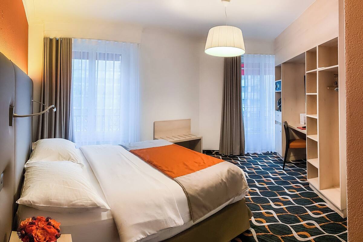 A hotel room with a bed, a desk, a chair, a wardrobe, and a hanging light fixture.
