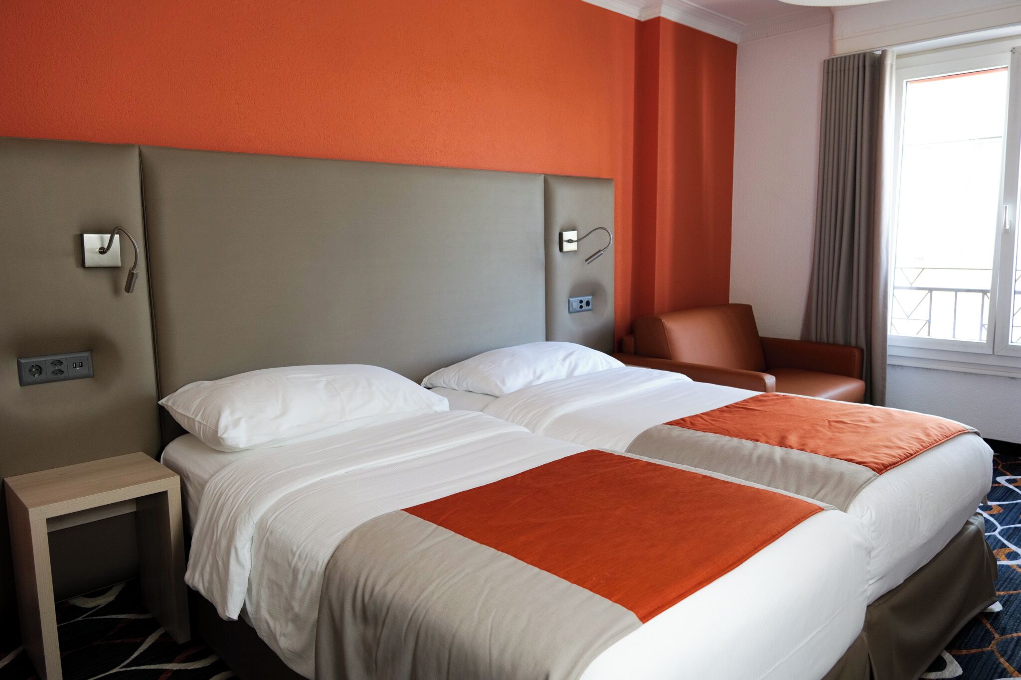 Ein Hotelzimmer mit zwei Betten, einem Nachttisch, einem Stuhl, einem Fenster mit Vorhängen und einer Wandsteuerung.