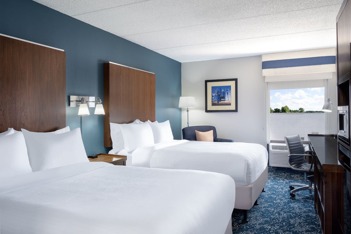 A hotel room with two beds, a desk, a chair, a lamp, and a picture on the wall.