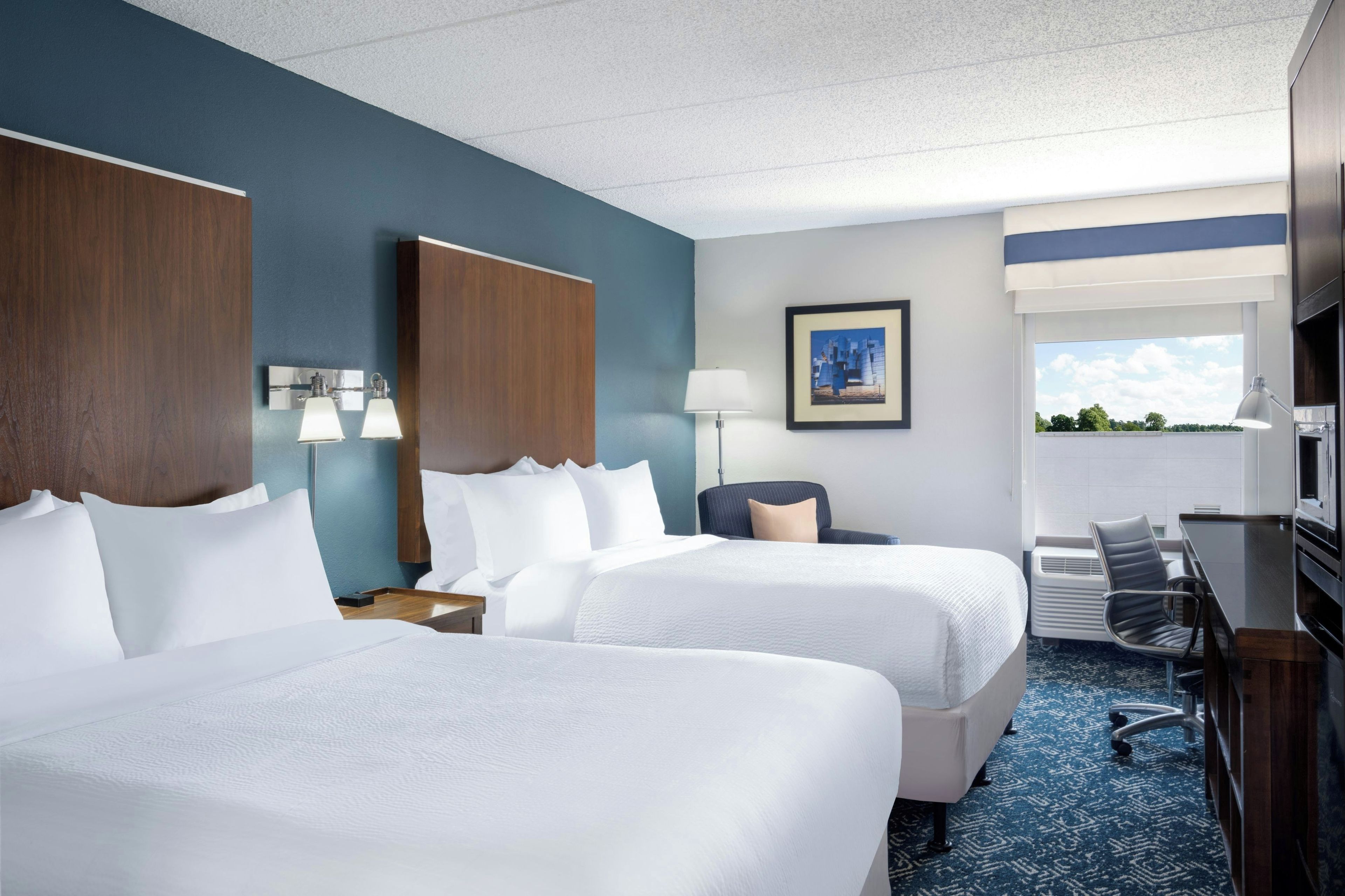 A hotel room with two beds, a desk, a chair, a lamp, a picture on the wall, and a window with a view.