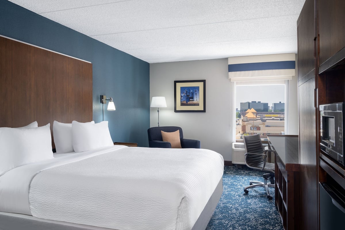 A hotel room with a large bed, a desk, a chair, a lamp, a framed picture on the wall, and a view of the city through a window.