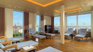 A spacious lounge area with wicker furniture, large windows offering city views, and a wooden floor. - Sheraton Carlton Hotel Nuernberg (Nuremberg)