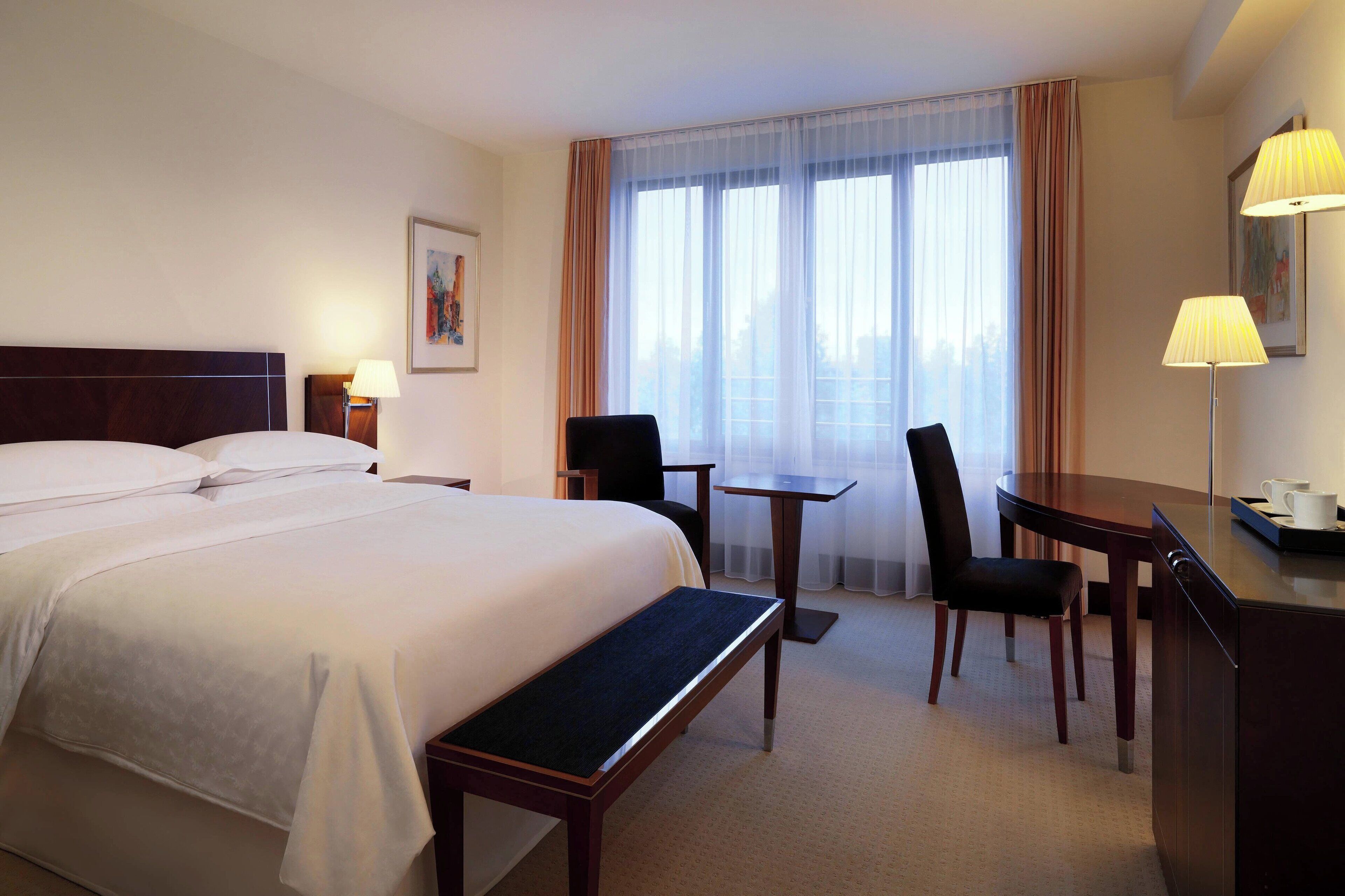 Quarto de hotel com uma cama grande, uma escrivaninha, duas cadeiras, uma mesinha e uma janela com cortinas.
