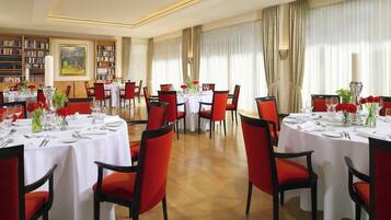A dining area with round tables, red chairs, and white tablecloths. There are bookshelves and a painting on the walls.