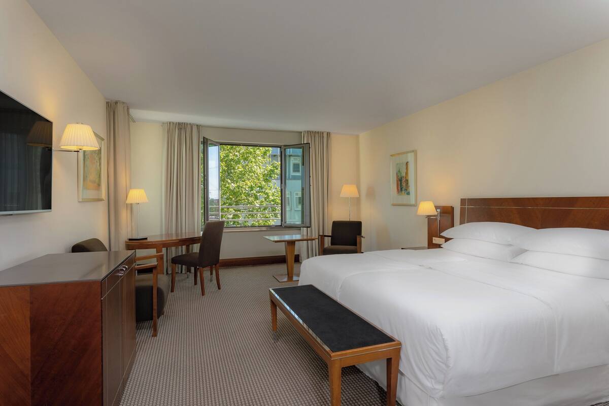 A hotel room with a large bed, a desk, two chairs, a bench, a TV, and a window with a view of greenery.