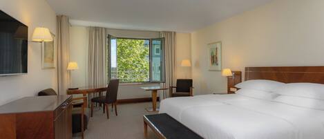 A hotel room with a large bed, a desk, two chairs, a bench, a TV, and a window with a view of greenery.