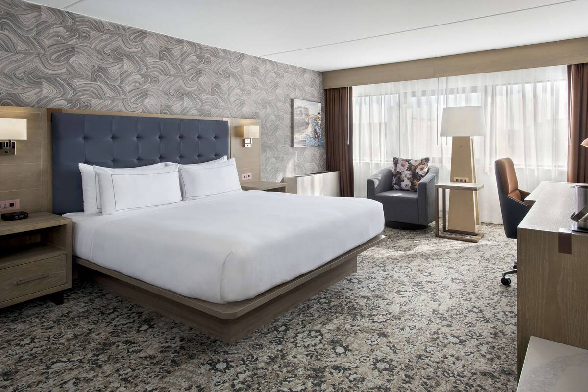 A hotel room with a large bed, a desk with a lamp, a chair, and a nightstand with a clock.