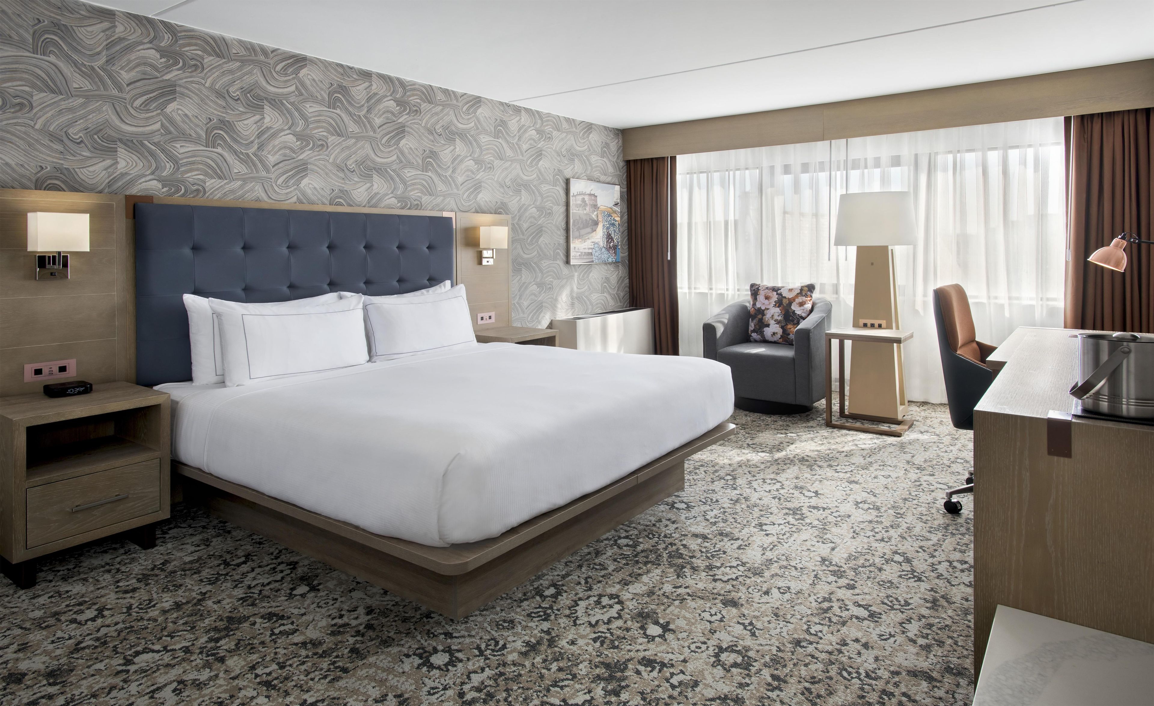 A hotel room with a large bed, a desk, a chair, and a view of the city through the window.