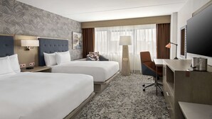 Habitación de hotel con dos camas, un escritorio, una silla, un televisor y un ventanal con cortinas.