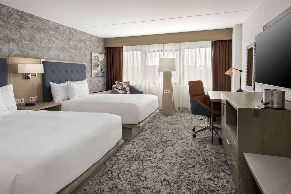 A hotel room with two beds, a desk, a chair, a TV, and a large window with curtains.