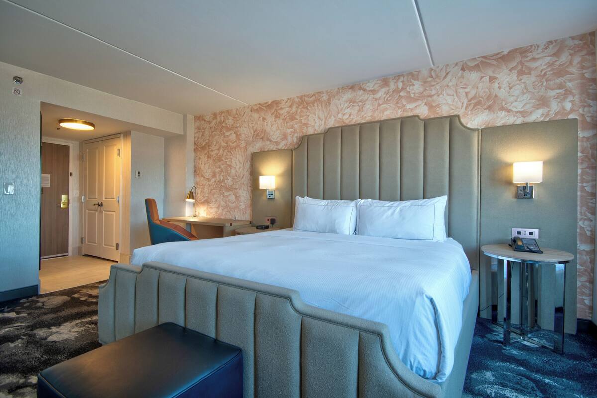 A hotel room with a large bed, a desk, a chair, a small table, and a bench.