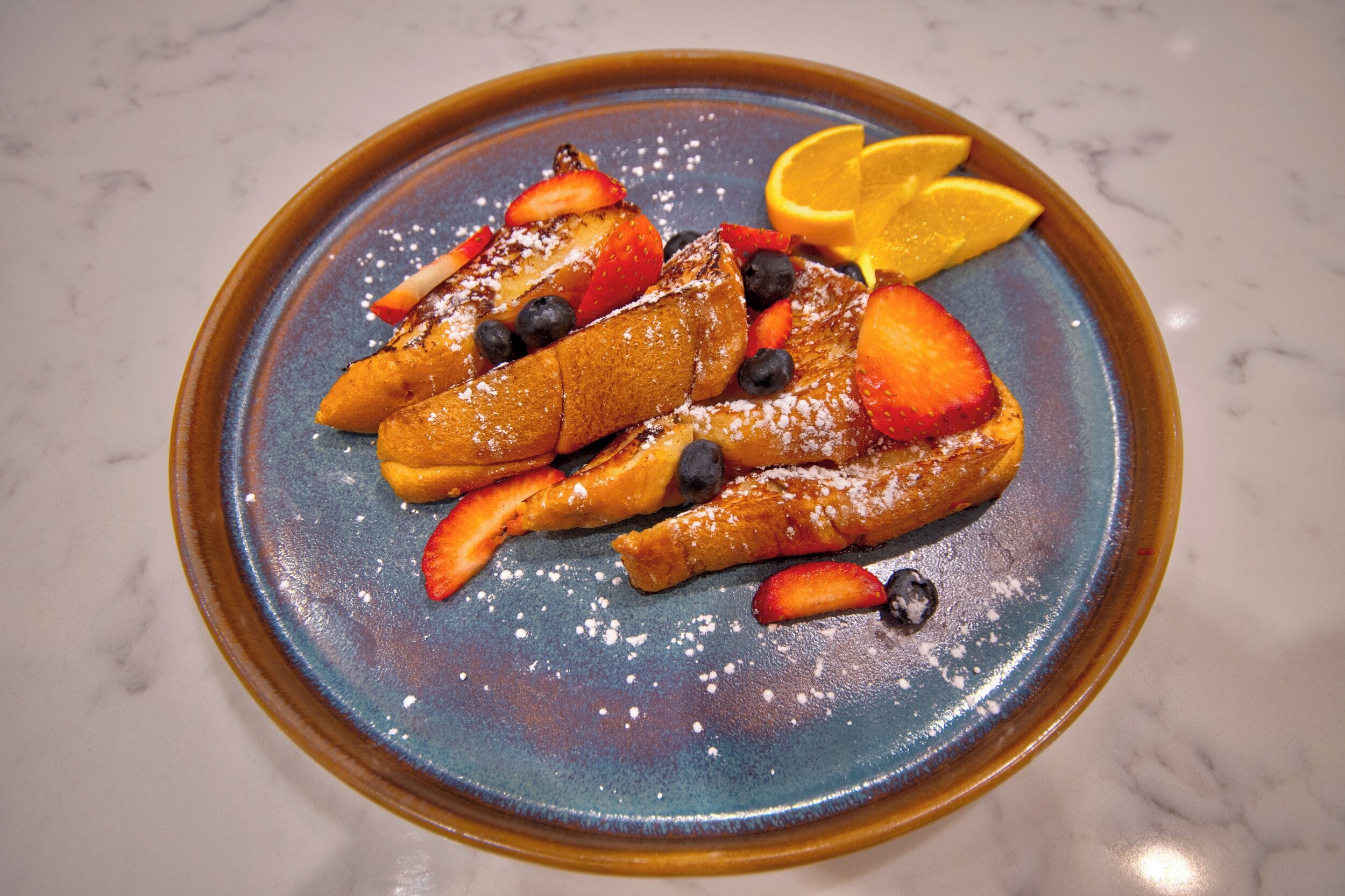 A plate of French toast with strawberries, blueberries, and orange slices on a blue ceramic plate.