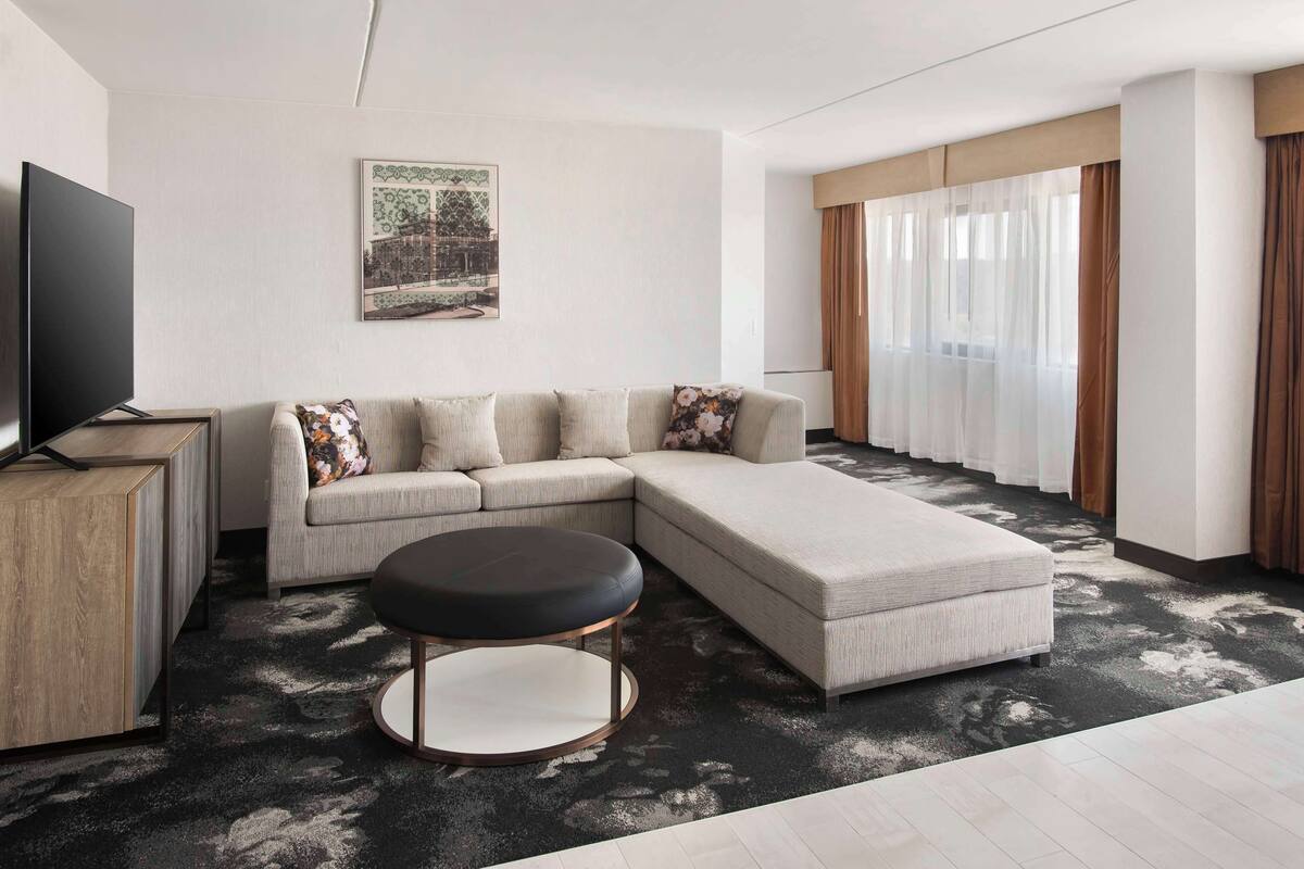 A modern living room with a sectional sofa, a round ottoman, a flat-screen TV, and a side table.
