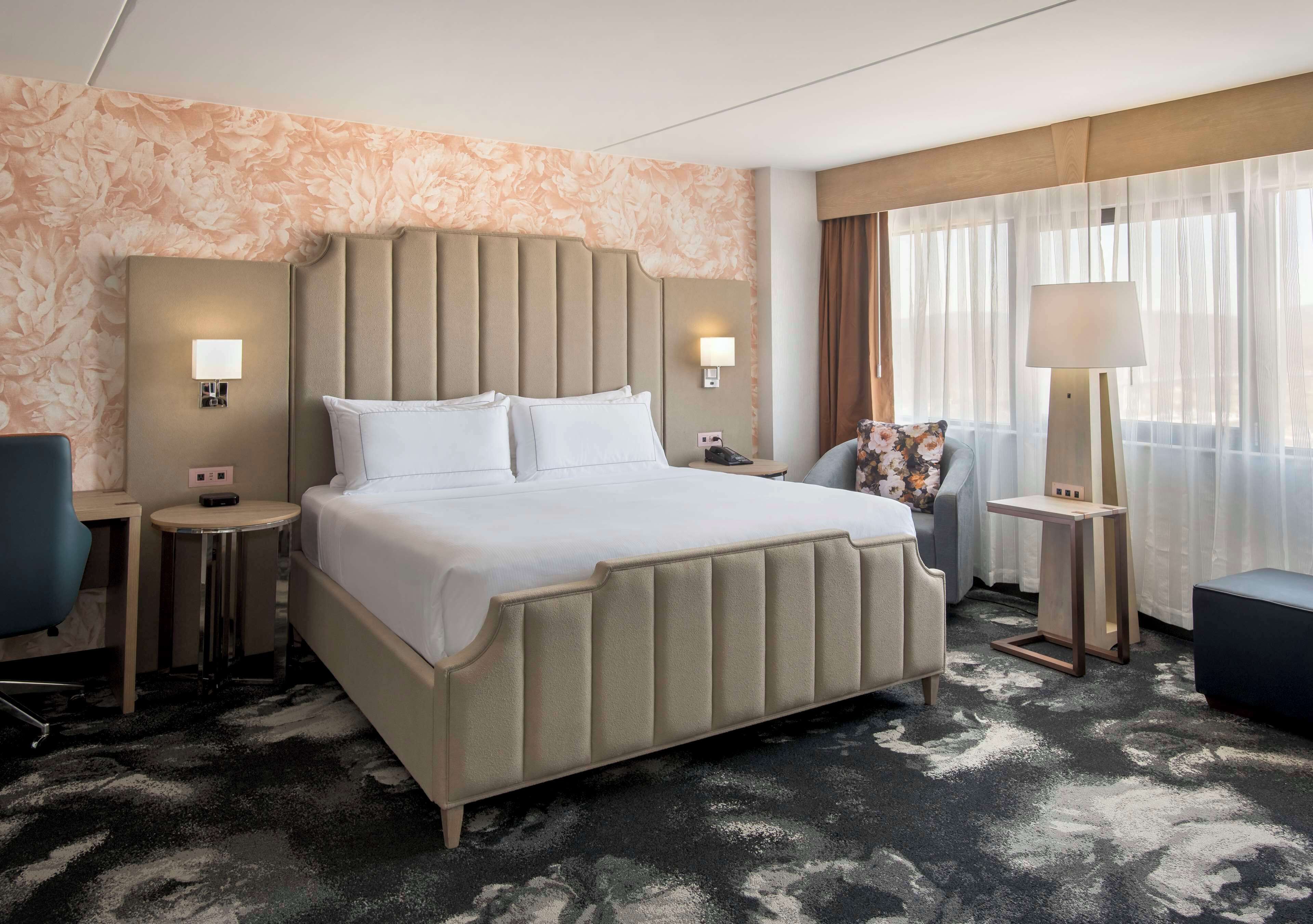 A hotel room with a large bed, two bedside tables, a chair, a lamp, and a window with curtains.
