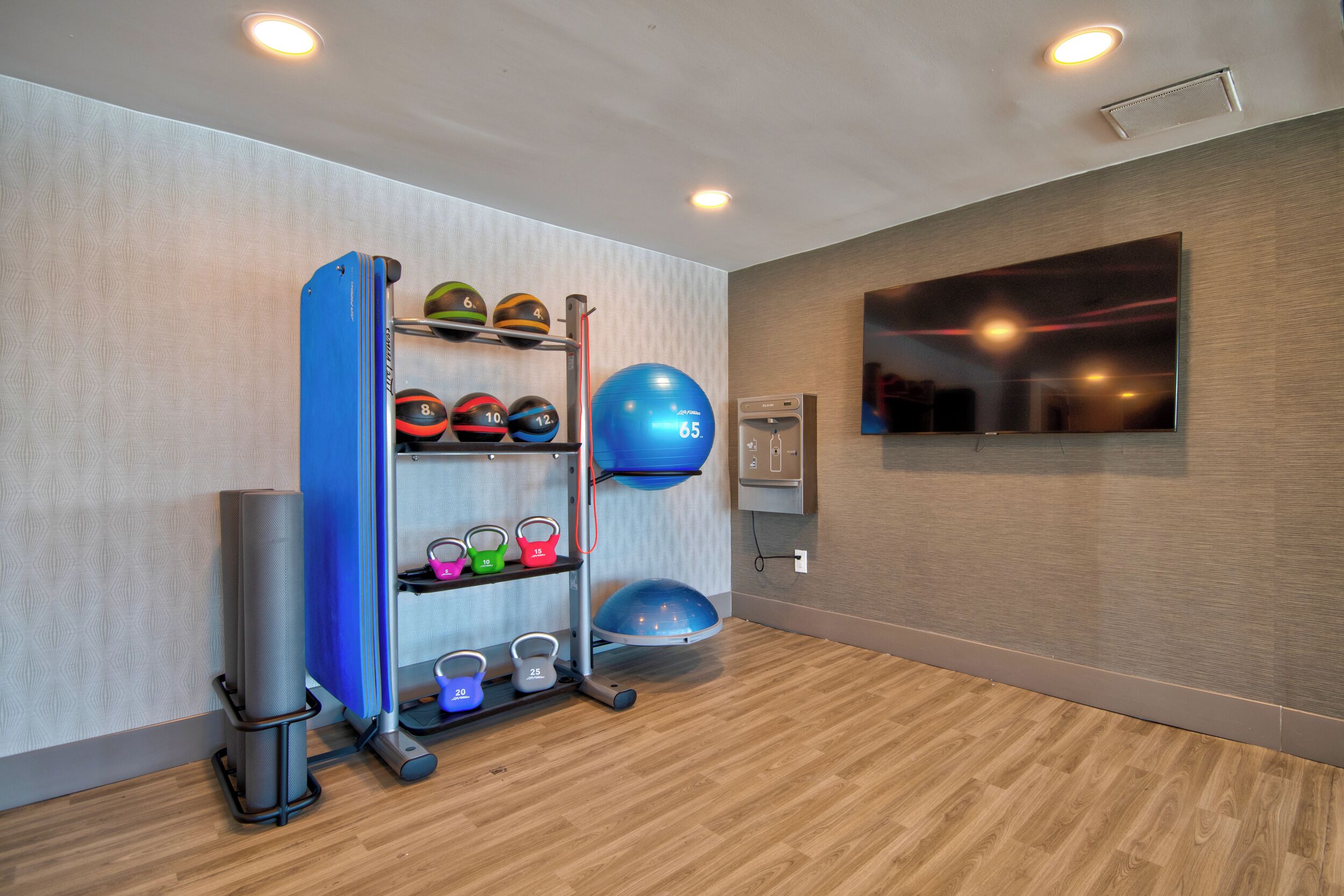A fitness room with exercise equipment, a wall-mounted TV, and a wooden floor.