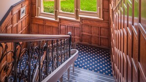 A wooden staircase with a patterned blue carpet, a chandelier, and a view of a green landscape through a window with floral curtains. - Bestwood Lodge (Nottingham)