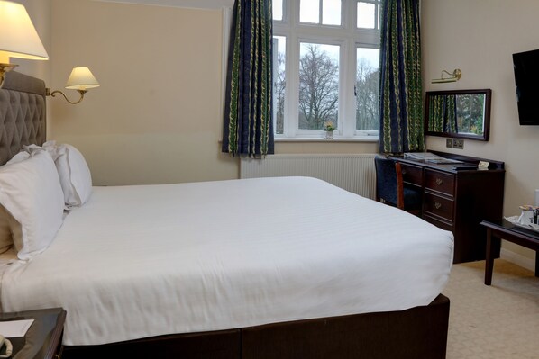 A neatly made bed with white linens, a bedside table with a lamp, a dresser with a mirror, and a window with curtains.