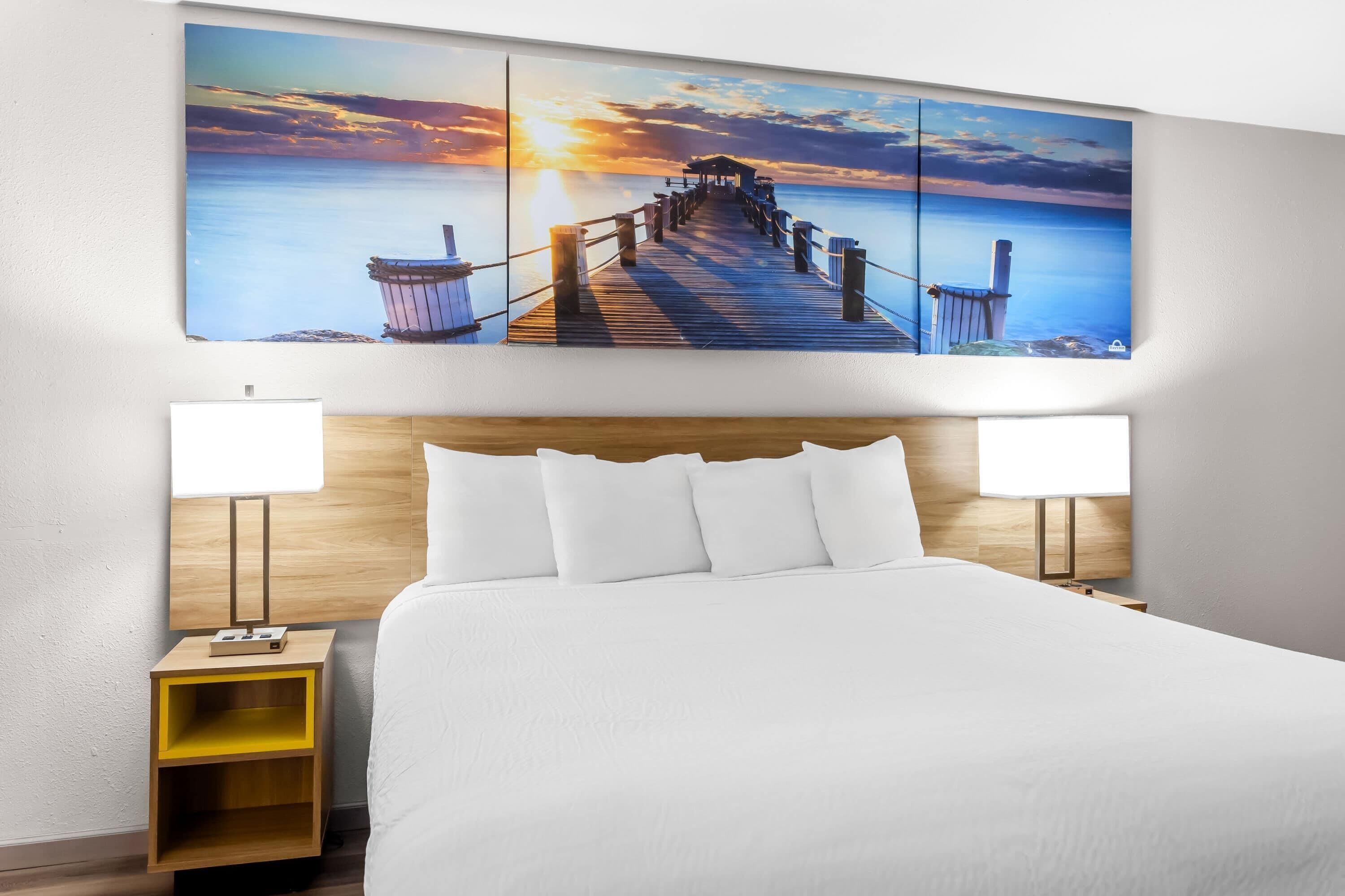 Una habitación de hotel con una cama grande, dos lámparas de noche, una mesita de noche de madera y un cuadro colgado en la pared que representa un muelle al atardecer.