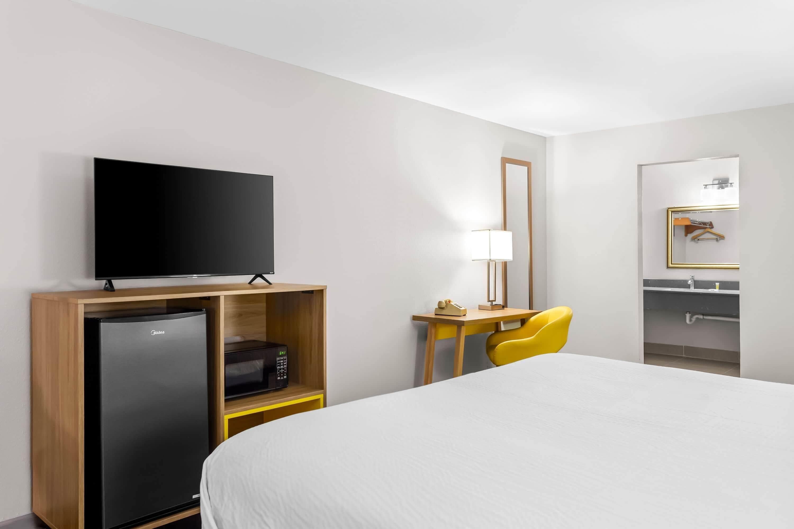 Et hotelværelse med seng, fjernsyn, køleskab, skrivebord med lampe, stol og spejl.