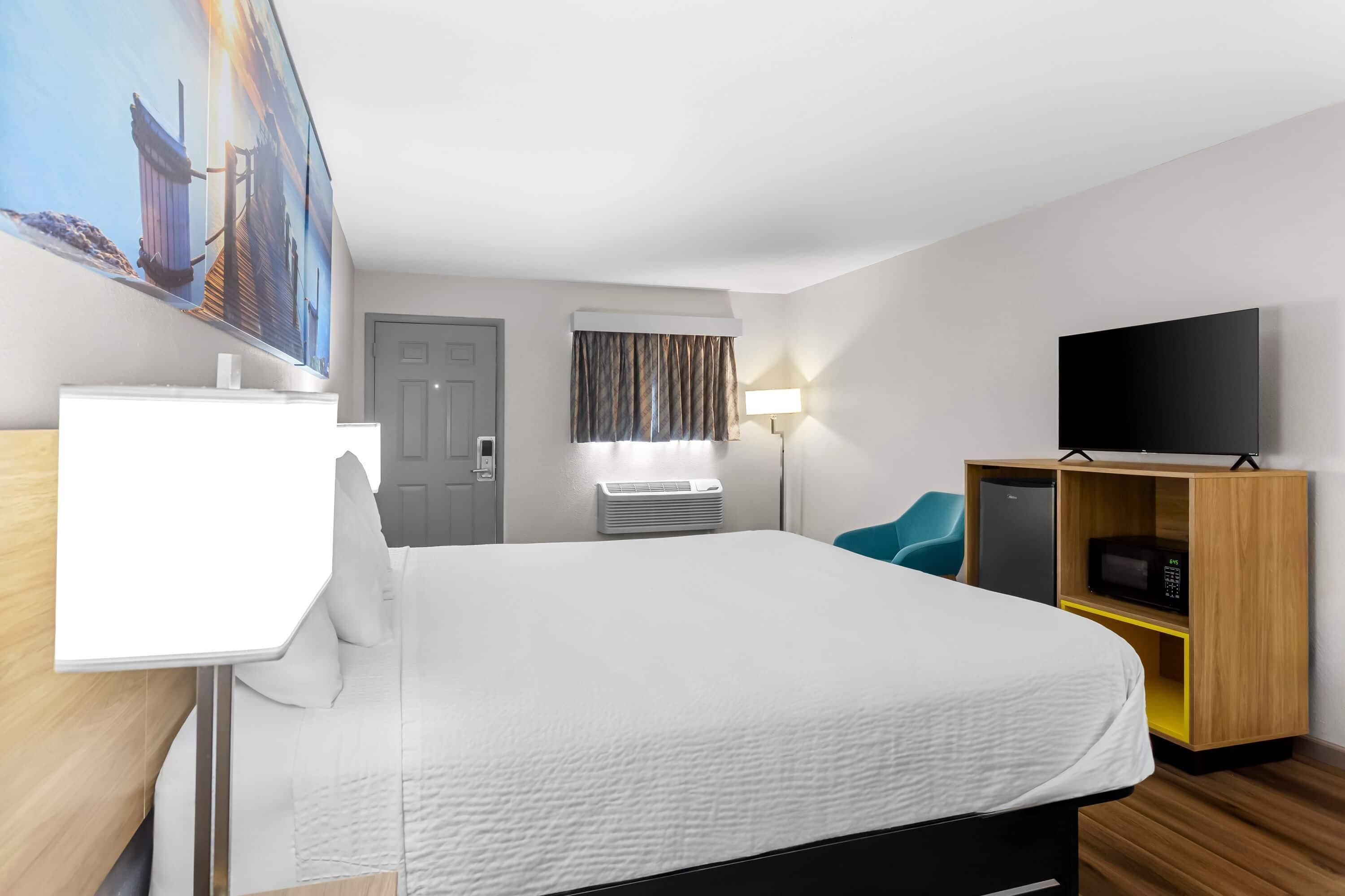 Ein Hotelzimmer mit Bett, Fernseher, Schreibtisch, Stuhl, Kleiderschrank und einem Bild an der Wand.