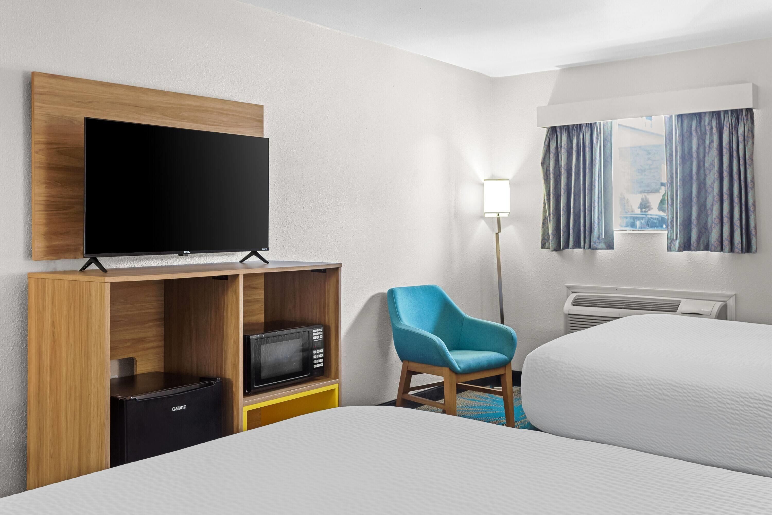A hotel room with a bed, a TV on a wooden shelf, a microwave, a chair, and a window with curtains.