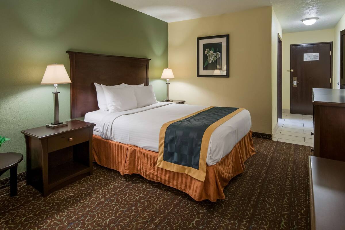 A hotel room with a bed, two bedside tables with lamps, a chair, a desk with a TV, and a door leading to another room.