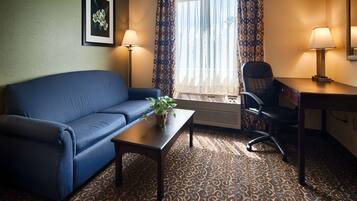 Habitación de hotel con un sofá azul, una mesita con una planta, un escritorio con una silla y un gran ventanal con cortinas.