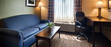 Habitación de hotel con un sofá azul, una mesita con una planta, un escritorio con una silla y un gran ventanal con cortinas.