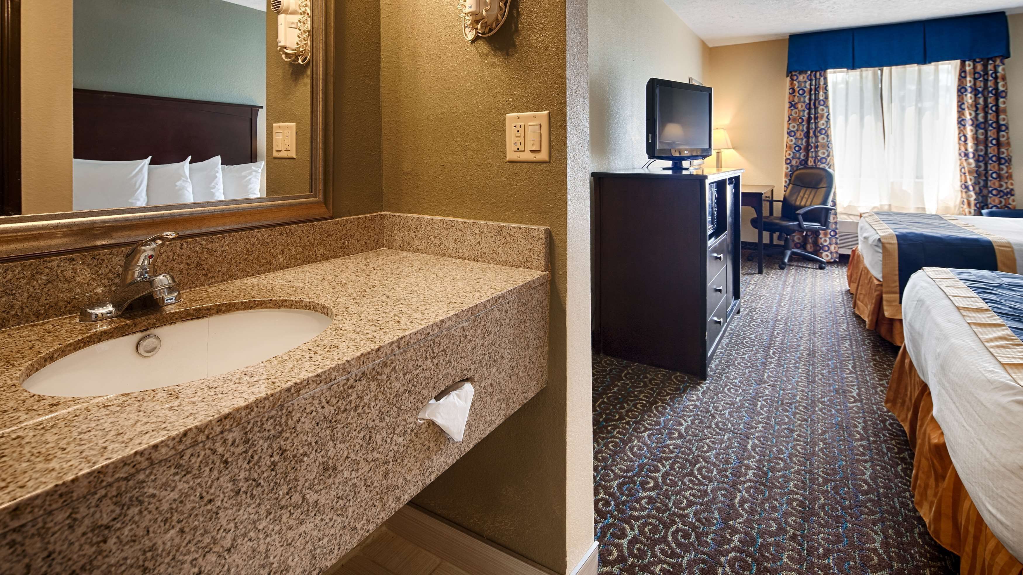 A hotel room with a granite countertop sink, a mirror, a television, a desk, and two beds with patterned bedding.