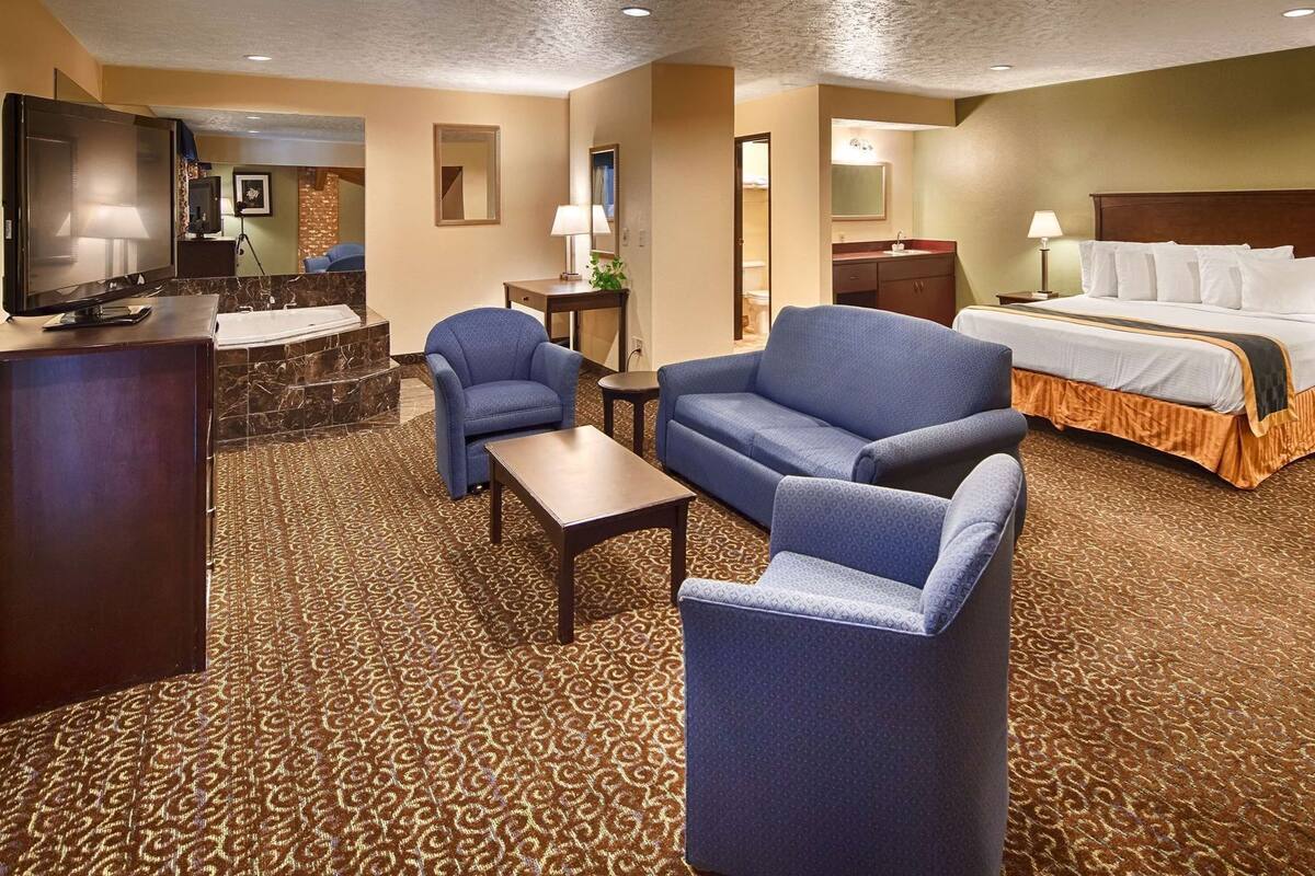 A hotel room with a bed, two sofas, a TV, a desk, and a bathroom visible through an open door.