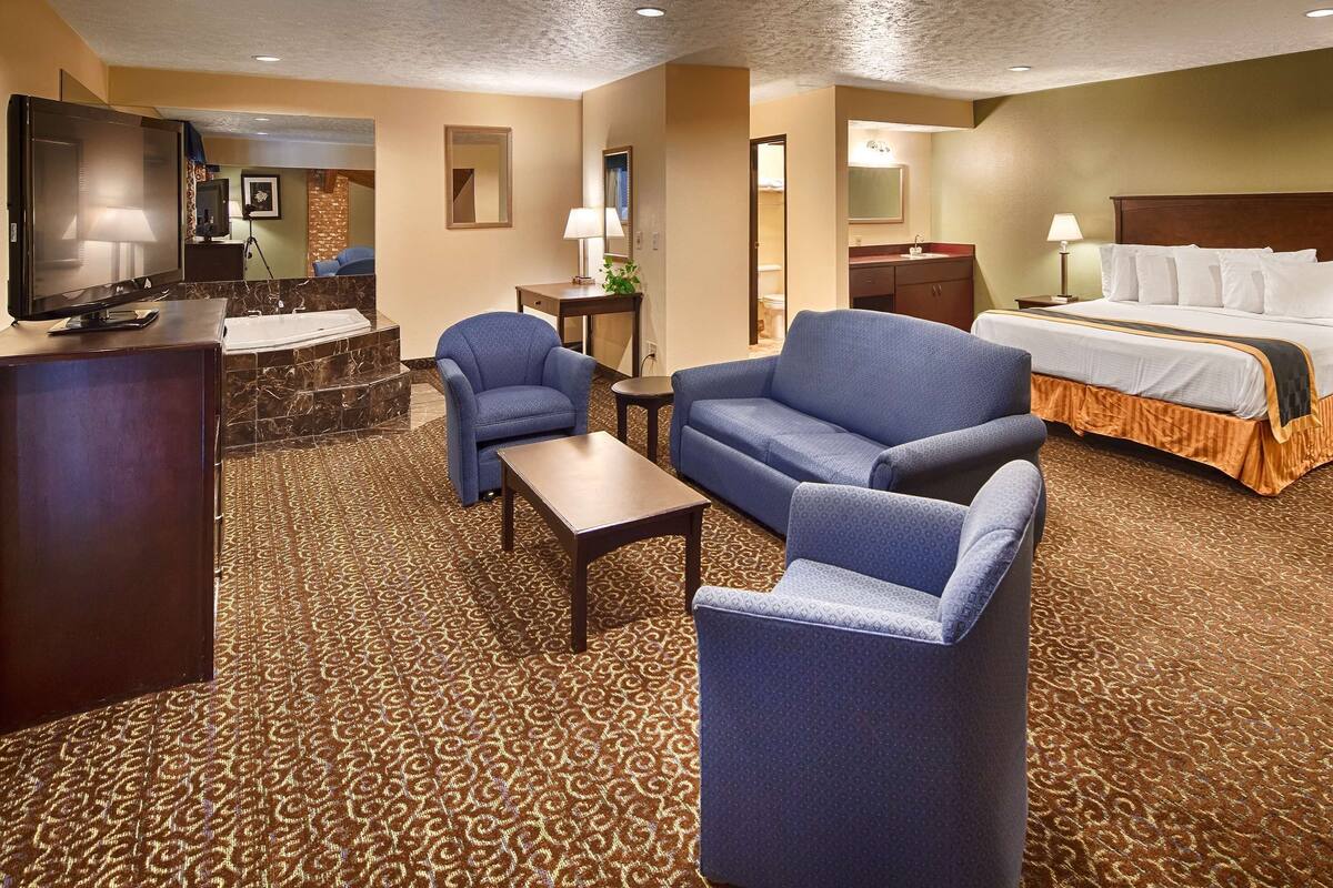 A hotel room with a large bed, two armchairs, a small table, a desk, a TV, and a bathroom visible through an open door.