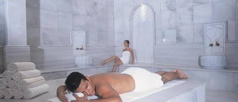 A spa room with marble walls, a large bathtub, and a towel stack.