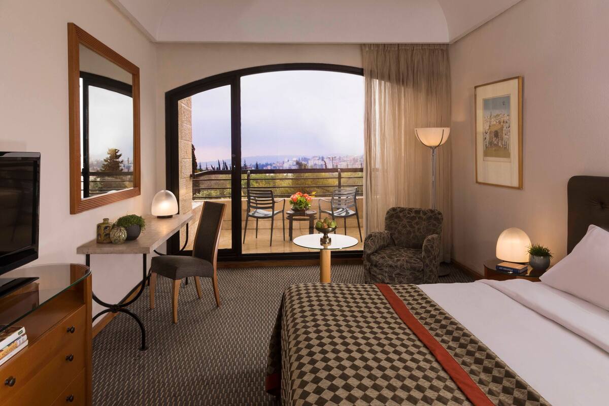 A hotel room with a bed, a desk, a chair, a small table with a plant, a lamp, and a view of the city through a large window.