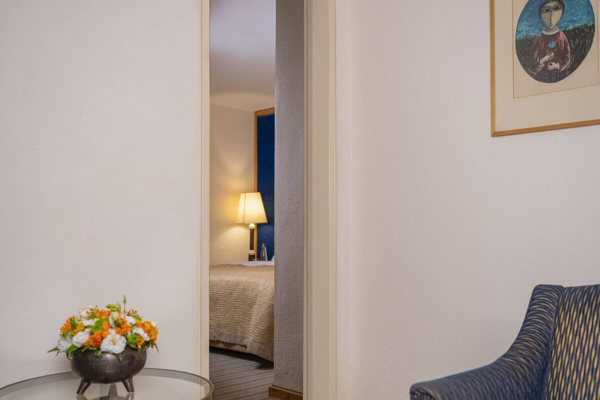 A hotel room with a striped carpet, a round table with a flower vase, a blue armchair, a bed with a blue headboard, and a framed picture on the wall.