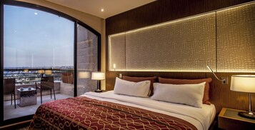 Habitación de hotel con una cama grande, vistas a la ciudad y un balcón con sillas.