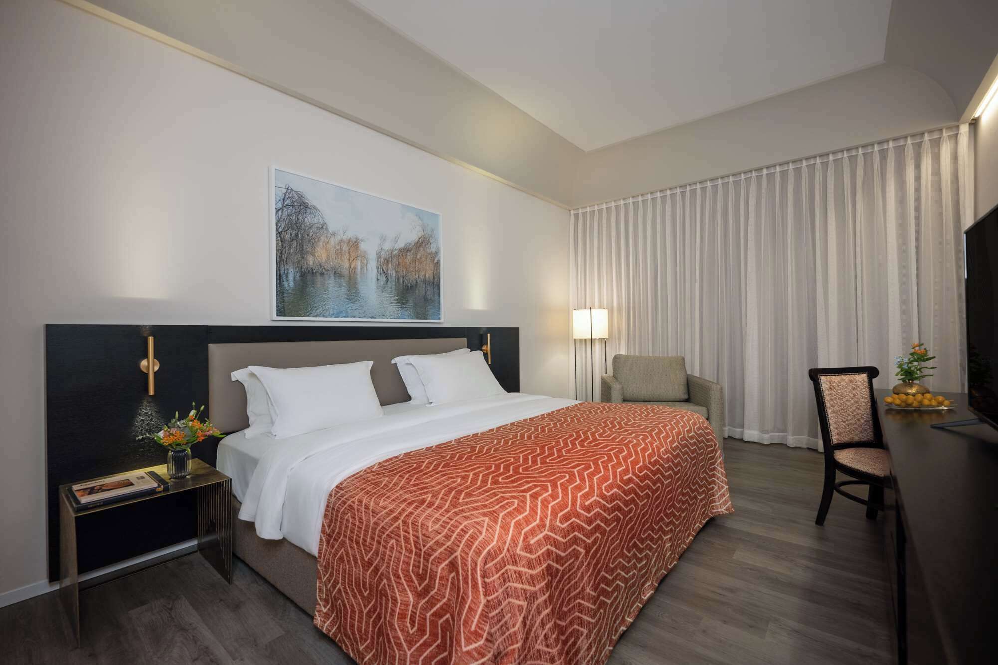 A hotel room with a bed, a chair, a desk, and a painting on the wall.