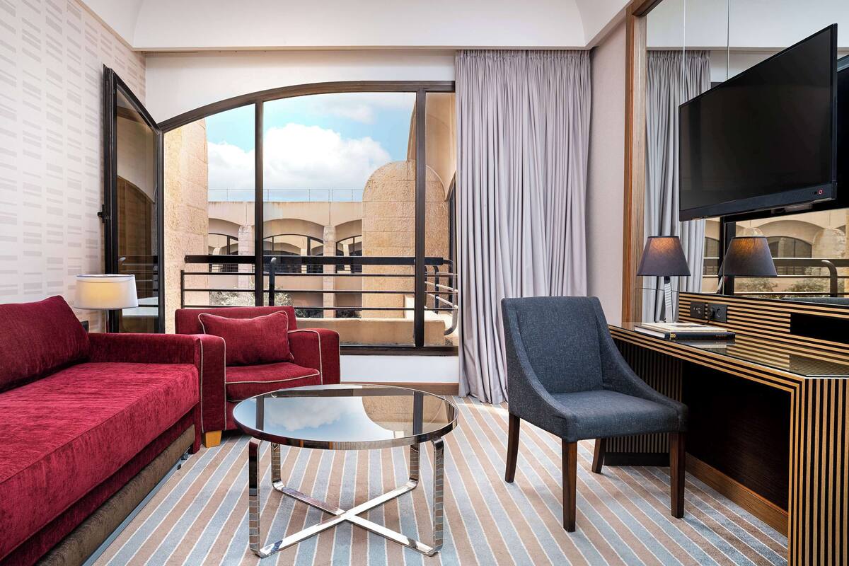 A hotel room with a red sofa, a blue armchair, a glass coffee table, a TV, and a balcony with a view of buildings.