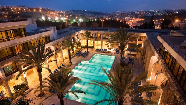 A rooftop swimming pool with lounge chairs and palm trees, surrounded by a building with large windows and a view of a cityscape at night. - Dan Jerusalem (Jerusalem)
