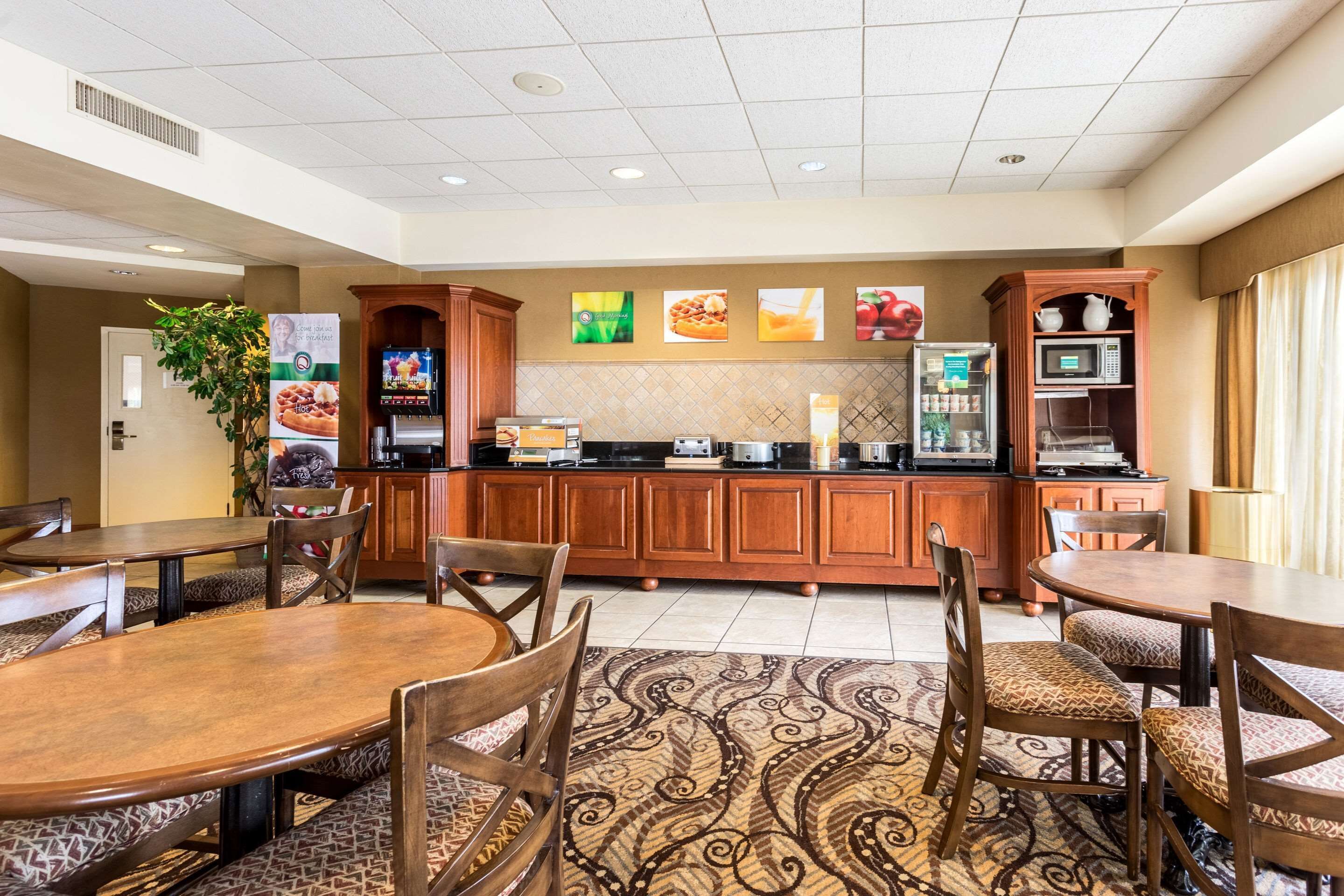 A hotel dining area with wooden tables and chairs, a patterned rug, and kitchen appliances.