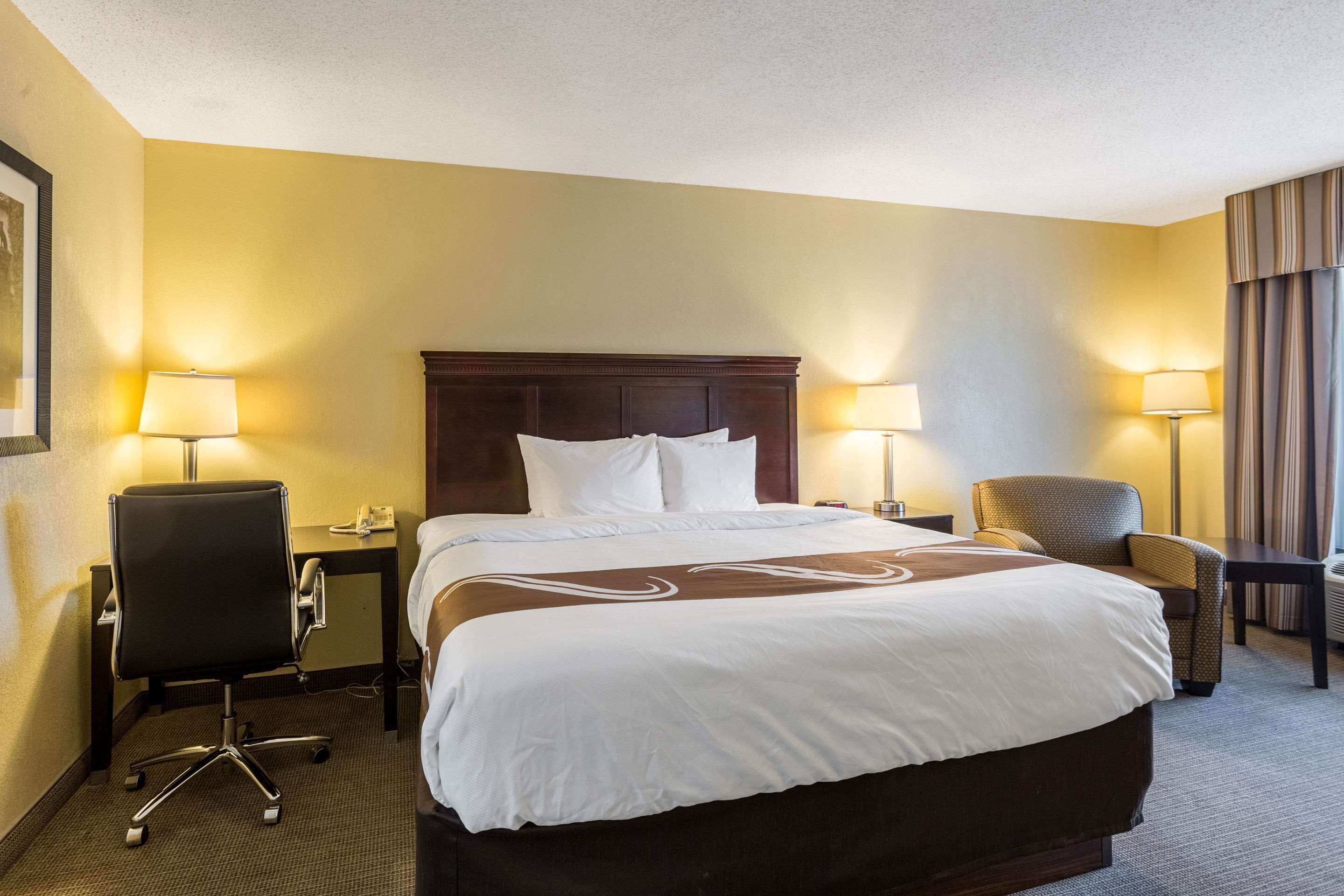 A hotel room with a large bed, two bedside lamps, a desk, a chair, and a window with curtains.