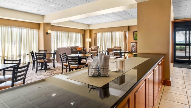 Un comedor de hotel con una barra larga, varias mesas y sillas. Hay cortinas en las ventanas, una lámpara y vistas al exterior.