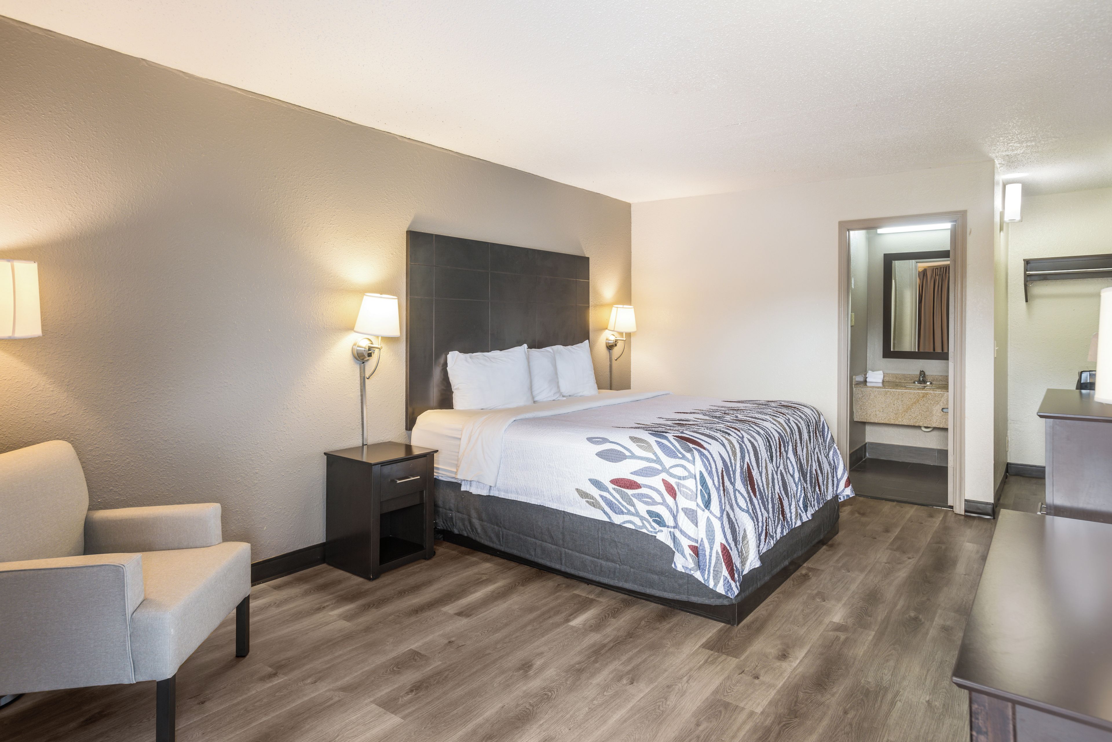 Habitación de hotel con cama, mesitas de noche, una silla y baño con espejo y lavamanos.