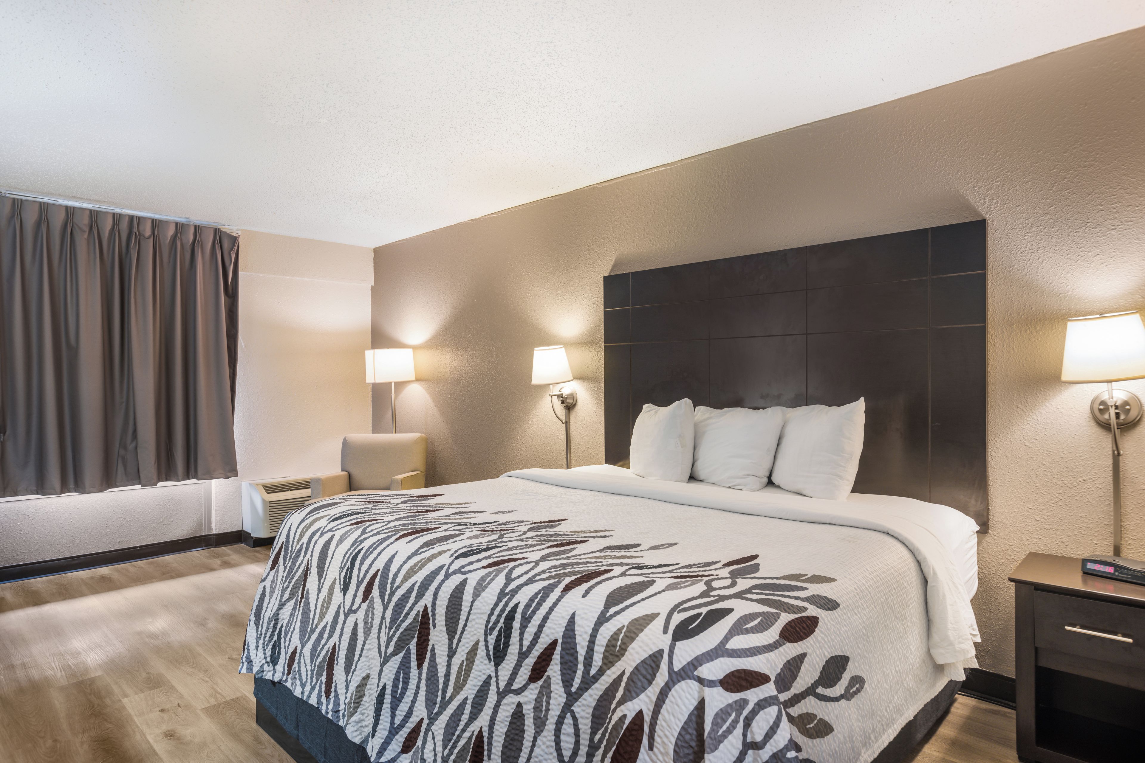 A hotel room with a large bed, two bedside lamps, a headboard, a nightstand, a window with curtains, and an air conditioning unit.