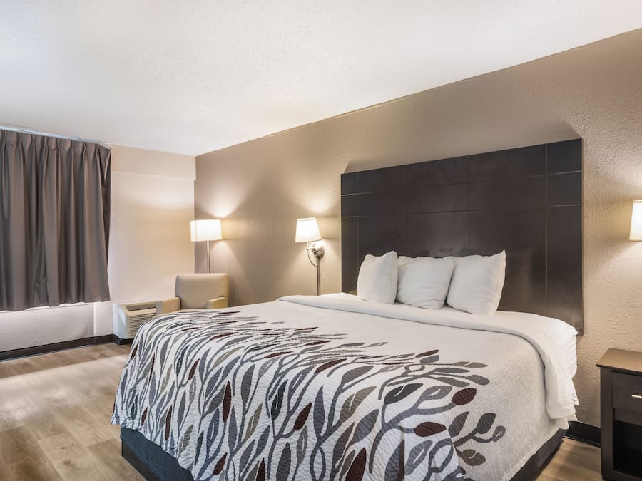 A hotel room with a large bed, two bedside lamps, a headboard, a nightstand, a window with curtains, and an air conditioning unit.